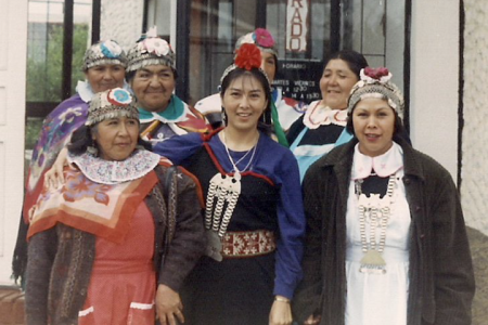 Seven Mapuche women of various ages wearing traditional clothing in shades of blue, purple, red, and black. They stand together in front of a building, posing for the camera.